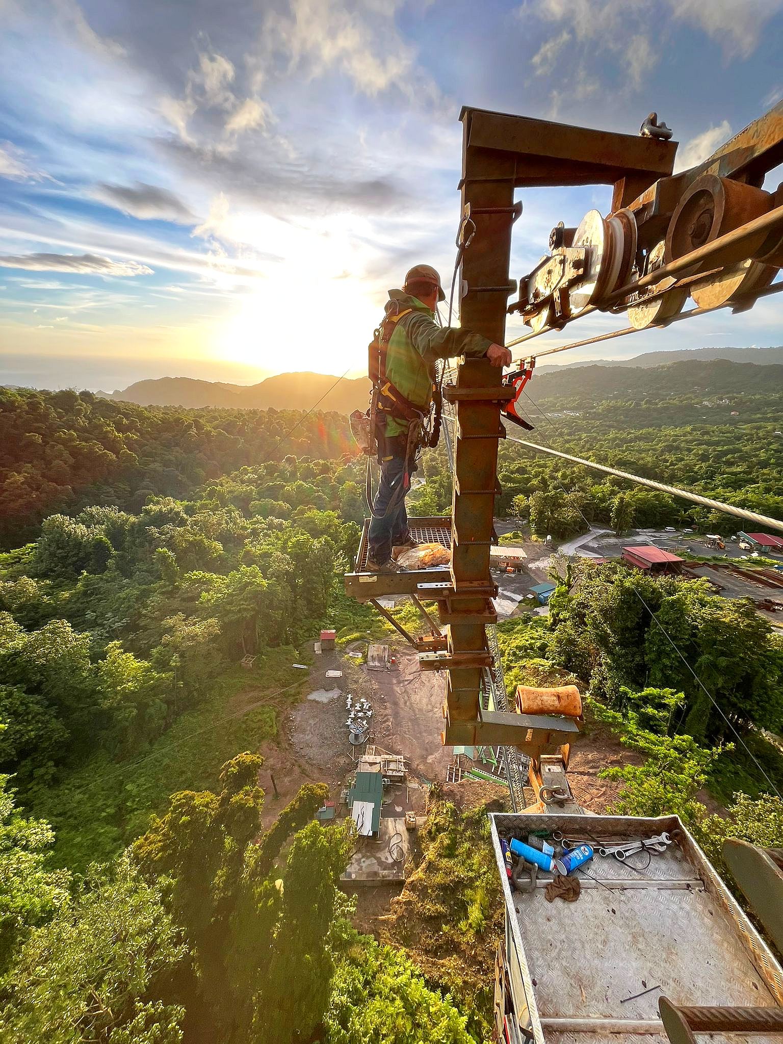 Monteur auf hohem Seilbahnpfeiler mit Baustelle und Tropenwald im Hintergrund.