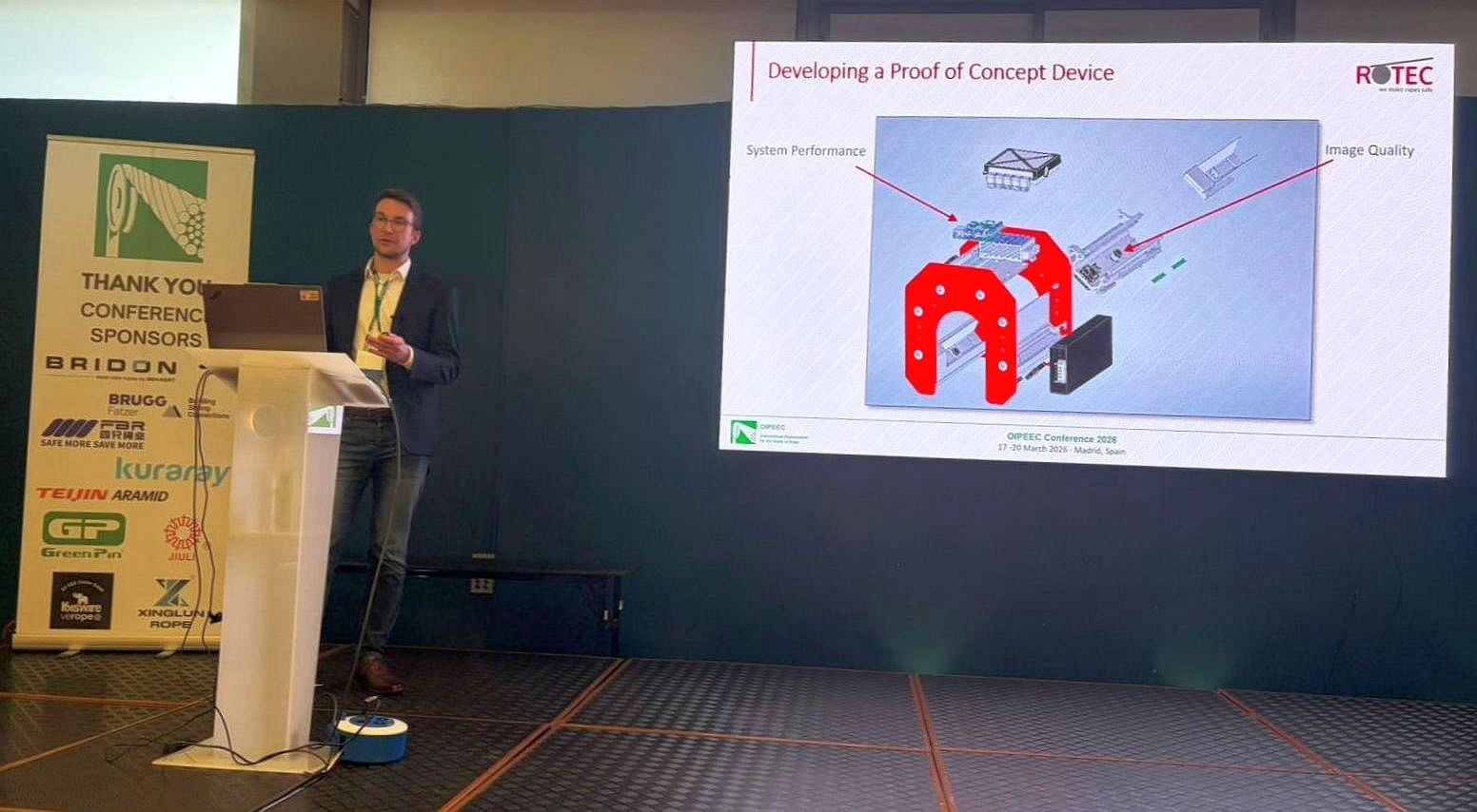 A man is speaking at a lectern on a stage next to a technical presentation slide.